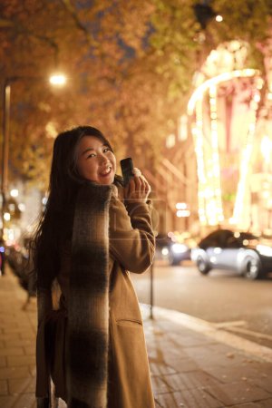 Téléchargez les photos : Asiatique fille à l'aide de téléphone mobile, debout dans la rue en prenant des photos de veilleuses. Femme chinoise aux cheveux longs, voyageant à travers l'Europe, marchant dans la ville. Concept tourisme, blogging, internet - en image libre de droit
