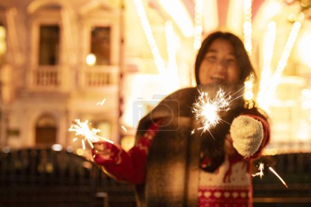 Foto de Mujer asiática feliz usando bufanda caliente sosteniendo chispas en las manos. Brillantes luces de Navidad bokeh en el fondo. - Imagen libre de derechos