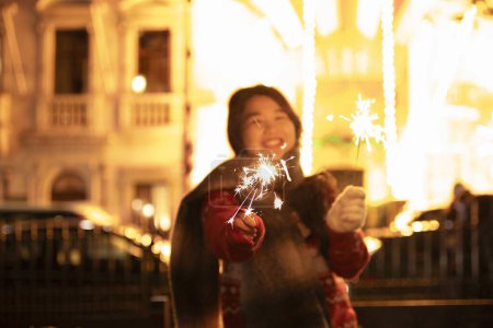 Téléchargez les photos : Heureuse femme asiatique portant une écharpe chaude tenant des étincelles dans les mains. Lumineux bokeh lumières de Noël sur fond. - en image libre de droit