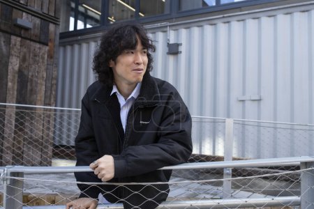 Téléchargez les photos : Élégant asiatique gars 25 ans avec de longs cheveux bouclés de bonne humeur portant une veste noire, debout à l'extérieur. Style de rue. - en image libre de droit