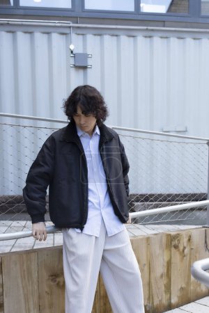 Téléchargez les photos : Portrait de jeune homme asiatique beau avec de longs cheveux bouclés debout à l'extérieur et regardant vers le bas. Style de rue. - en image libre de droit