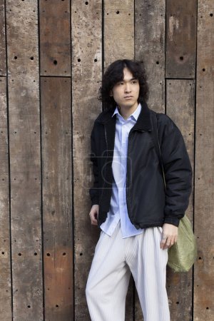 Photo for Stylish young Japanese young man with long curly hair. Serious, calm face, hard look. Wooden boards on background. Street style photo. - Royalty Free Image