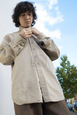 Schöner, modisch gekleideter asiatischer Mann mit lockigem Haar predigt sich selbst und befestigt Knopf an seiner Jacke. Konzept für Modeblogger, Stylisten, Designer. Blauer Himmel Hintergrund