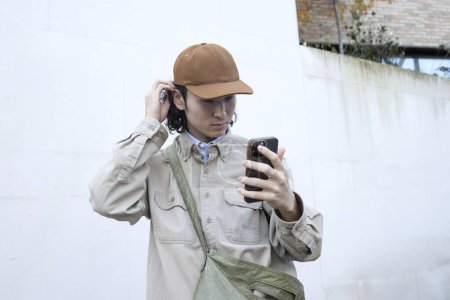 Schöner cool gekleideter asiatischer Mann predigt sich selbst, schaut in die Kamera seines Telefons, steckt sich die Haare hinter das Ohr und trägt eine Baseballmütze. Konzept für Modeblogger, Stil, Designer. 