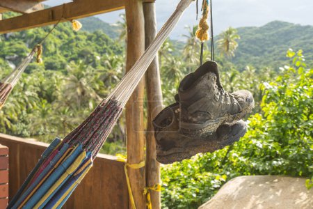Photo for Muddy and dirty Hiking Boots hanging in the air by their shoe laces next to a hammock at Tayrona national park in Columbia, South America. Green rainforest jungle in the background - Royalty Free Image