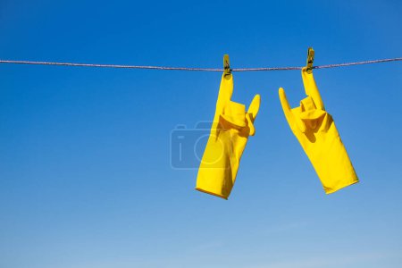 Photo for Yellow rubber protective gloves for work or cleaning hanging on a rope with a clothespin against a blue sky background. The gloves are hung by the index fingers and show rock gesture - Royalty Free Image