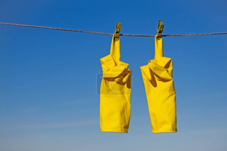 Téléchargez les photos : Gants de protection en caoutchouc jaune pour le travail ou le nettoyage suspendus à une corde avec une pince à linge sur un fond bleu ciel. Les gants sont accrochés par les doigts et montrent un geste du majeur vers le haut - en image libre de droit