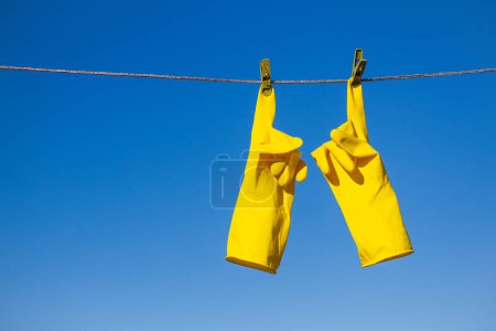 Téléchargez les photos : Gants de protection en caoutchouc jaune pour le travail ou le nettoyage suspendus à une corde avec une pince à linge sur un fond bleu ciel. Les gants sont suspendus par les index et montrent un geste pointu - en image libre de droit