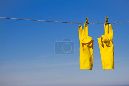 Téléchargez les photos : Gants de protection en caoutchouc jaune pour le travail ou le nettoyage suspendus à une corde avec une pince à linge sur un fond bleu ciel. Les gants sont accrochés par les doigts du milieu et montrent un geste de victoire - en image libre de droit