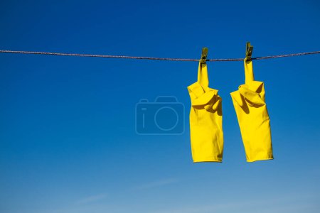 Photo for Yellow rubber protective gloves for work or cleaning hanging on a rope with a clothespins against a blue sky background. The gloves are hung by the fingers and show middle finger up gesture - Royalty Free Image