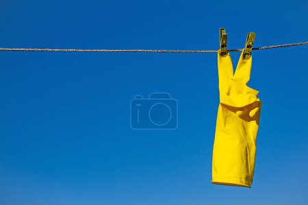 Foto de Guantes protectores de goma amarilla para trabajar o limpiar colgados de una cuerda con pinzas de ropa sobre un fondo azul del cielo. Los guantes están colgados de los dedos medios y muestran el gesto de victoria - Imagen libre de derechos