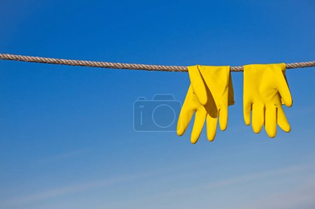 Téléchargez les photos : Gants de protection en caoutchouc jaune pour le travail ou le nettoyage accrochés à une corde et séchés dans les rayons du soleil sur un fond bleu ciel - en image libre de droit