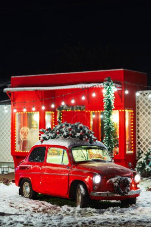 Téléchargez les photos : Une voiture rouge avec un sapin de Noël sur le toit dans le contexte d'un magasin rouge. vieille voiture rouge avec des décorations de nouvelle année et de la neige dans la rue. Zone photo de Noël avec une voiture pour les séances photo de famille - en image libre de droit