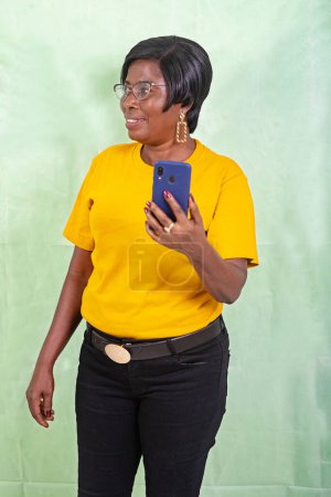 Photo pour Une belle femme mûre dans un t-shirt jaune debout sur un fond vert regardant loin tenant un téléphone portable tout en souriant. - image libre de droit