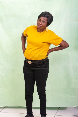 Foto de Una hermosa mujer con una camiseta amarilla de pie sobre un fondo verde que sufre de dolor de espalda. - Imagen libre de derechos
