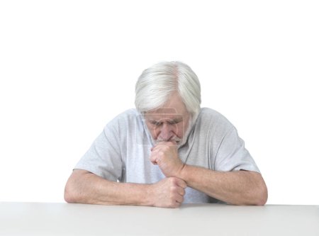 Foto de Horizontal shot of a white haired old man sitting at a table leaning his head on his hands in grief, worry or prayer.  Isolated on white.  Lots of copy space. - Imagen libre de derechos