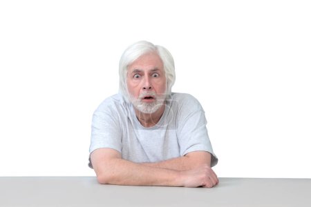 Photo for Horizontal shot of an old man sitting at a table with his arms crossed looking very surprised.  Isolated on white.  Lots of copy space. - Royalty Free Image