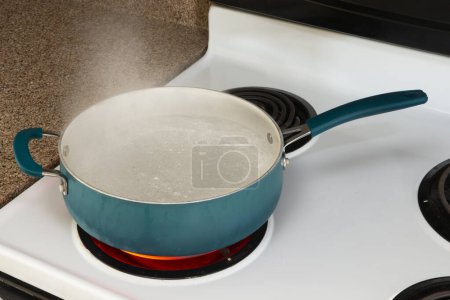 Disparo horizontal mirando hacia abajo en una olla azul en una estufa blanca en un quemador caliente rojo sosteniendo agua hirviendo. 