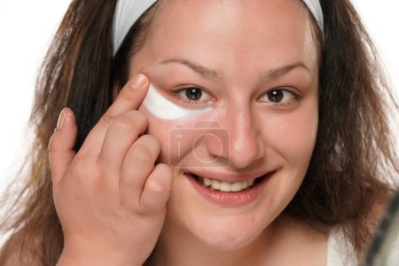 Foto de Retrato de una joven mujer sonriente gordita aplicando producto cosmético bajo sus ojos sobre un fondo blanco. - Imagen libre de derechos