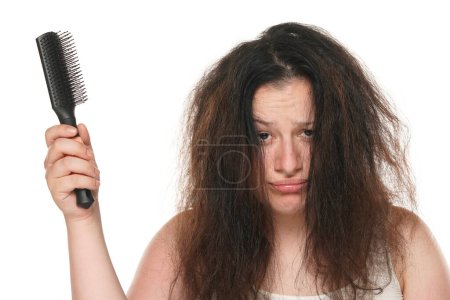 Téléchargez les photos : Portrait d'une jeune femme potelée nerveuse aux cheveux longs et salissants tenant une brosse à cheveux sur fond de studio blanc. - en image libre de droit