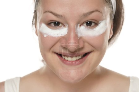 Téléchargez les photos : Portrait d'une jeune femme souriante potelée avec un produit cosmétique sous les yeux sur fond blanc. - en image libre de droit