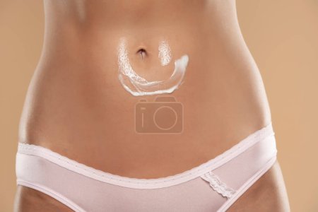 Téléchargez les photos : Close-up partial view of woman with body cream on belly on a beige studio background - en image libre de droit