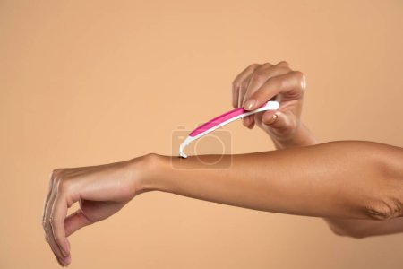 Téléchargez les photos : Une femme se rase les bras avec un rasoir rose jetable sur fond de studio beige - en image libre de droit