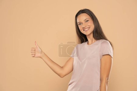 Foto de Portrait of young happy woman showing thumbs up on beige studio background - Imagen libre de derechos
