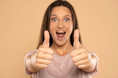 Téléchargez les photos : Portrait of young happy woman showing thumbs up on beige studio background - en image libre de droit