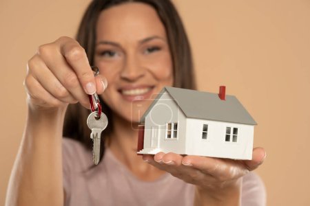 Foto de Young woman smiling and holding house sample model and keys  isolated over beige studio background, Real estate and home insurance concept - Imagen libre de derechos