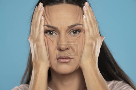 Photo for Headshot of a young woman crumples the skin of her face with her hands on a blue background - Royalty Free Image
