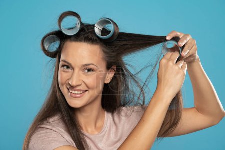 Photo for Young woman putting curlers in her hair on a blue studio background - Royalty Free Image