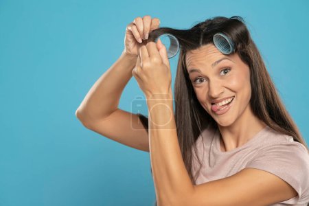 Foto de Young woman putting curlers in her hair on a blue studio background - Imagen libre de derechos