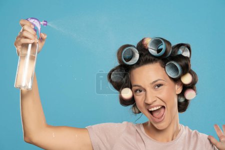 Téléchargez les photos : Happy femme pulvérise de la laque sur ses bigoudis sur un fond de studio bleu - en image libre de droit