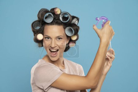 Photo for Happy woman sprays hairspray on her hair curlers on a blue studio background - Royalty Free Image