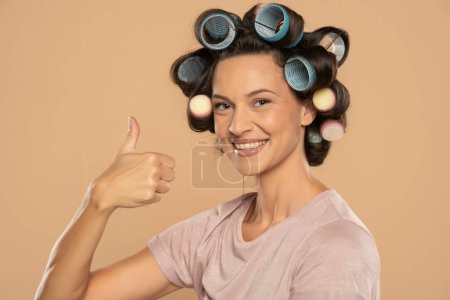 Téléchargez les photos : Belle femme souriante avec des boucles de cheveux montrant pouces vers le haut sur un fond studio beige - en image libre de droit