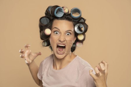 Foto de Hermosa mujer loca enojada con rizadores de pelo posando sobre un fondo de estudio beige - Imagen libre de derechos