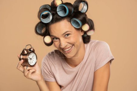 Téléchargez les photos : Belle femme souriante avec des bigoudis tenant une horloge sur un fond studio beige - en image libre de droit