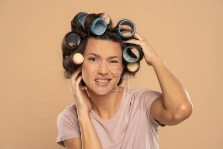Photo for Beautiful nervous woman with hair curlers scratching her head on a beige studio  background - Royalty Free Image