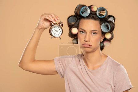 Téléchargez les photos : Belle femme ennuyée avec des bigoudis tenant une horloge sur un fond studio beige - en image libre de droit