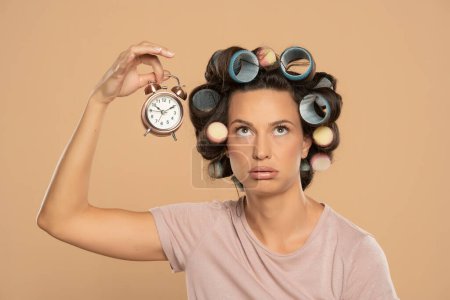 Téléchargez les photos : Beautiful bored woman with hair curlers holding a clock on a beige studio  background - en image libre de droit