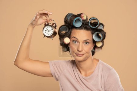 Téléchargez les photos : Belle femme ennuyée avec des bigoudis tenant une horloge sur un fond studio beige - en image libre de droit