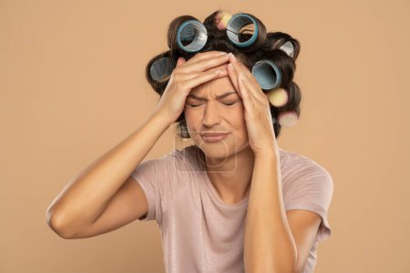 Téléchargez les photos : Beautiful woman with hair curlers and headache posing on a beige studio  background - en image libre de droit