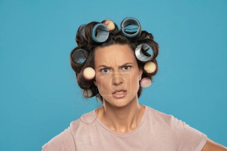 Photo for Beautiful angry woman with hair curlers posing on a blue studio  background - Royalty Free Image