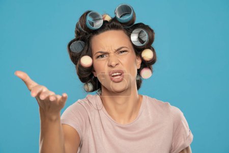 Téléchargez les photos : Belle femme en colère avec des bigoudis se disputent sur un fond de studio bleu - en image libre de droit