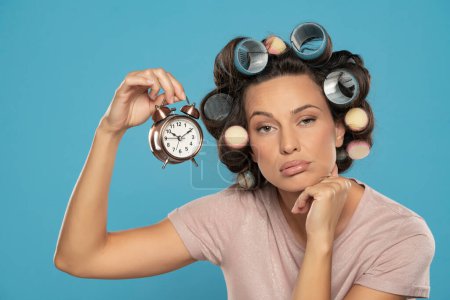 Photo for Beautiful bored woman with hair curlers holding a clock on a blue studio  background - Royalty Free Image