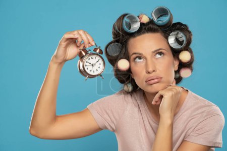 Téléchargez les photos : Belle femme ennuyée avec des bigoudis tenant une horloge sur un fond de studio bleu - en image libre de droit