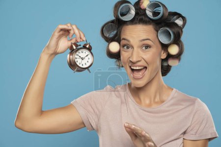 Foto de Hermosa mujer feliz con rizadores de pelo sosteniendo un reloj sobre un fondo de estudio azul - Imagen libre de derechos