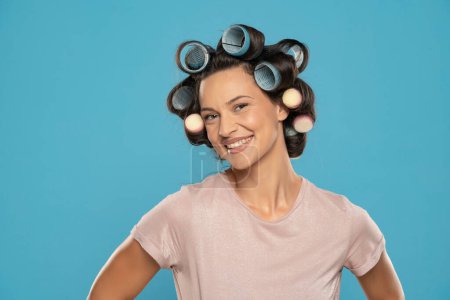 Photo for Beautiful smiling woman with hair curlers posing on a blue studio  background - Royalty Free Image