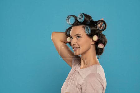 Photo for Beautiful smiling woman with hair curlers posing on a blue studio  background - Royalty Free Image